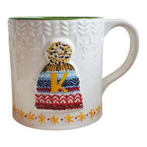 May include: White ceramic mug with a green rim and a large handle. The mug features a colorful, patterned design of a winter hat with the letter "K" on it. The base of the mug has yellow star-shaped accents.