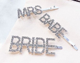 Könnte beinhalten: Drei silberne Haarklammern mit den Wörtern "MRS", "BRIDE" und "BRIDE", die mit Strasssteinen verziert sind.