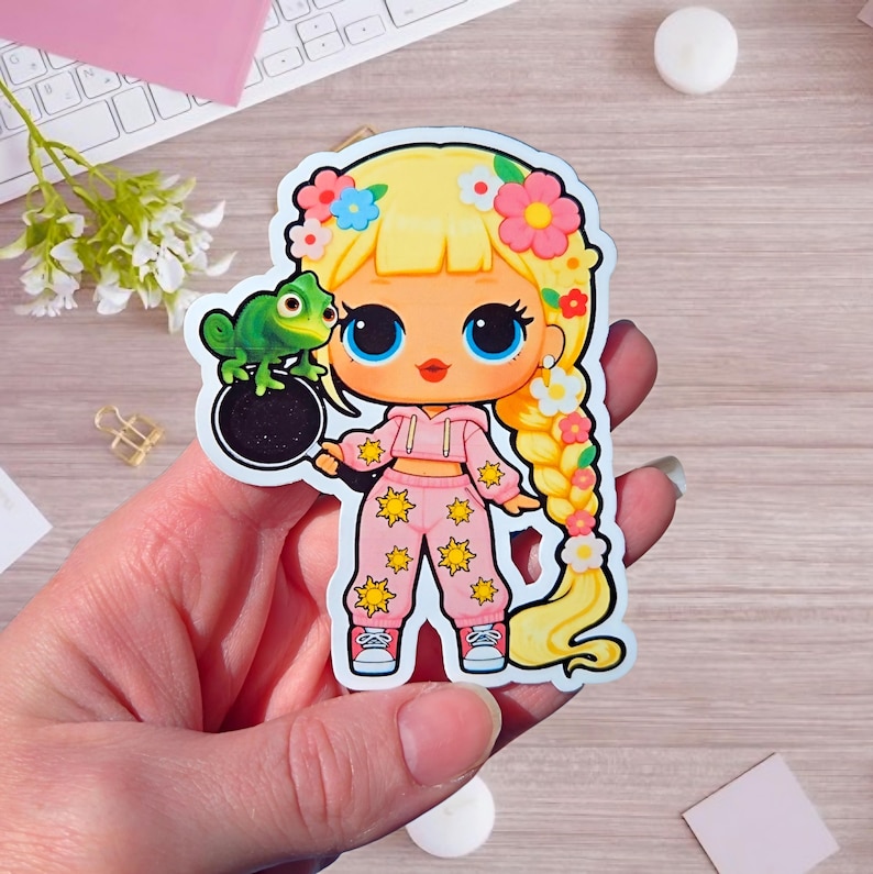 May include: A die-cut sticker featuring a cartoon character with long blonde braided hair adorned with colorful flowers. The character wears a pink hooded crop top and matching pants with yellow sun patterns, holding a black frying pan with a green chameleon.