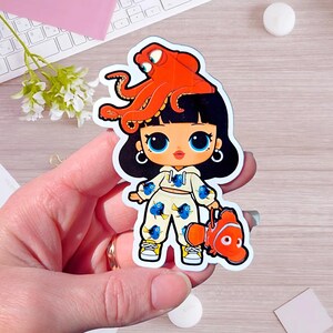 May include: A die-cut sticker featuring a cartoon character with black hair, large blue eyes, and a cream-colored hoodie and pants set patterned with blue fish. An orange octopus sits on the character's head, and an orange clownfish is held in hand.