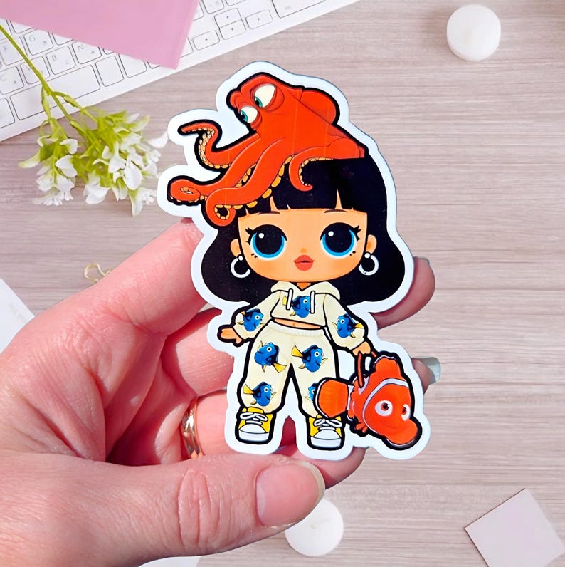 May include: A die-cut sticker featuring a cartoon character with black hair, large blue eyes, and a cream-colored hoodie and pants set patterned with blue fish. An orange octopus sits on the character's head, and an orange clownfish is held in hand.