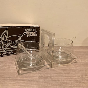 May include: Clear acrylic condiment server-2 set with two glass bowls, each with a small spoon. The set includes a box with the text "Acrylic Condiment Server-2". The bowls sit on a clear acrylic base.
