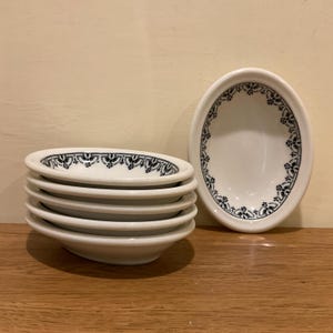 May include: A stack of six white, oval-shaped serving dishes with a black floral border. One dish is standing upright. The dishes are on a wooden surface.