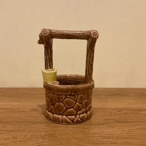 May include: A brown ceramic wishing well planter featuring a textured stone-like base and a faux wood grain support structure. A small, pale yellow bucket is attached to the side of the well rim, completing the miniature garden ornament design.
