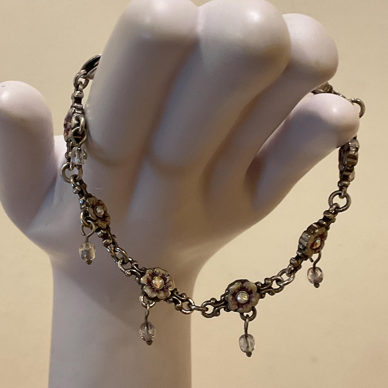 Peut inclure: Un bracelet &agrave; cha&icirc;ne de couleur argent&eacute;e compos&eacute; de maillons m&eacute;talliques en forme de fleurs r&eacute;p&eacute;t&eacute;es, avec de petites perles de cristal transparentes et facett&eacute;es suspendues &agrave; chaque fleur, reli&eacute;es par des anneaux m&eacute;talliques circulaires.