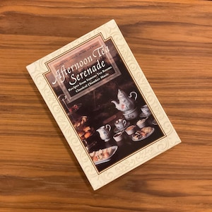 以下が含まれることがあります： シャロン・オコナー著『Afternoon Tea Serenade』というタイトルの長方形の本。白い縁取りにはエンボス加工のスクロール模様が施されている。表紙には、ティーポット、カップ、食べ物が載った皿が並ぶティーセットの暗い写真が印刷されている。