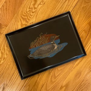 Puede incluir: Bandeja rectangular negra con una ilustración decorativa de un ave somorgujo sobre un fondo de agua azul. El somorgujo tiene un cuerpo moteado y está rodeado de líneas doradas y juncos marrones.