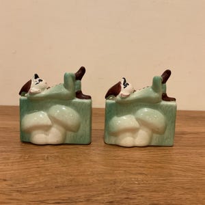 May include: A pair of ceramic figurines featuring a figure in a green outfit and brown hat reclining on a light green mushroom base. The figures have white skin, dark hair, and are positioned horizontally with legs raised, resting on a textured surface.