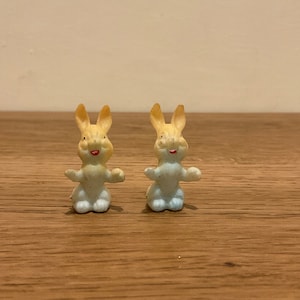 May include: Two small, white plastic bunny figurines with yellow ears and red painted smiles. The bunnies are sitting upright with their arms outstretched.