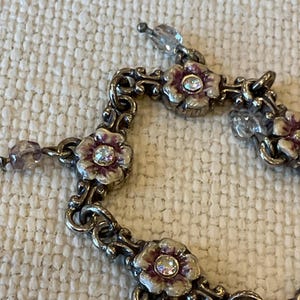 Peut inclure: Un bracelet en m&eacute;tal argent&eacute; compos&eacute; d'une s&eacute;rie de maillons floraux interconnect&eacute;s. Chaque fleur pr&eacute;sente des p&eacute;tales en &eacute;mail blanc et violet avec un strass transparent au centre. Des perles facett&eacute;es translucides sont fix&eacute;es entre les maillons.