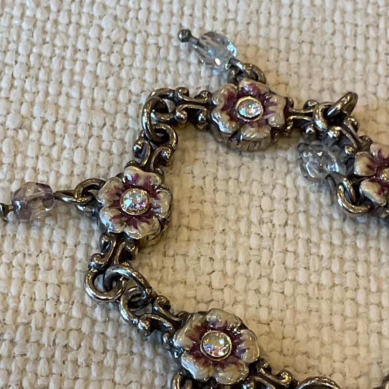 Peut inclure: Un bracelet en m&eacute;tal argent&eacute; compos&eacute; d'une s&eacute;rie de maillons floraux interconnect&eacute;s. Chaque fleur pr&eacute;sente des p&eacute;tales en &eacute;mail blanc et violet avec un strass transparent au centre. Des perles facett&eacute;es translucides sont fix&eacute;es entre les maillons.