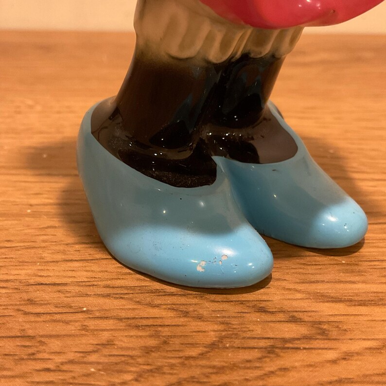 Op de afbeelding: Een keramiek beeldje van een meisje dat een roze jurk en blauwe schoenen draagt. Het beeldje staat met haar armen omhoog en haar hoofd naar achteren gekanteld. Het beeldje is realistisch geschilderd.