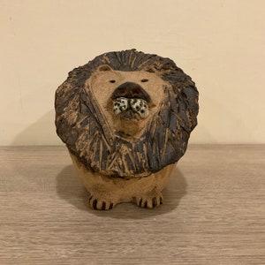May include: A ceramic lion figurine with a brown and black mane. The lion has a textured surface and a detailed face with black eyes and a white nose.