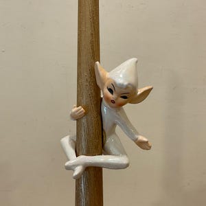 May include: A ceramic elf figurine with a glossy white finish, pointed ears, and a matching white hat, depicted clinging to a textured, light brown wooden pole. The elf features painted facial details including dark eyes and red lips.