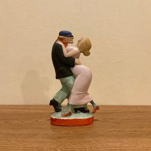 May include: A ceramic figurine of a couple dancing. The man is wearing a blue hat, black jacket, and plaid pants. The woman is wearing a pink dress. The figurine is painted in bright colors and has a whimsical style.