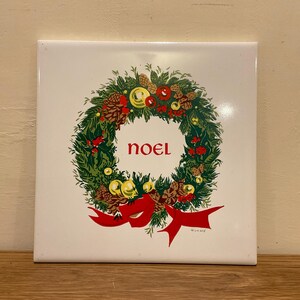 May include: Square white tile with a Christmas wreath design. The wreath is green with red berries, pinecones, and a red bow. The word "NOEL" is in red in the center of the wreath.