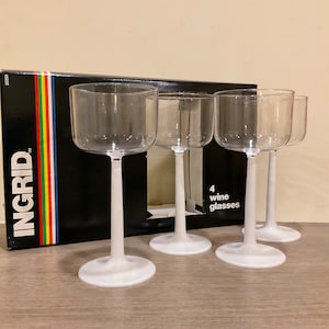 May include: Four clear wine glasses with white stems and bases are displayed in front of a black box with the word "INGRID" and a colorful stripe design. The box also indicates that there are four wine glasses.