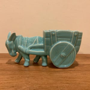 May include: A light blue ceramic planter shaped like a donkey pulling a wooden-style cart. The donkey is harnessed to the cart, which features a slatted design and a single large wheel on the side. The entire piece has a glossy, smooth finish.