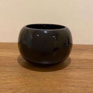 May include: A glossy black ceramic bowl with a rounded shape. The bowl has a wide opening and sits on a wooden surface. The smooth, reflective surface of the bowl catches the light, creating highlights.