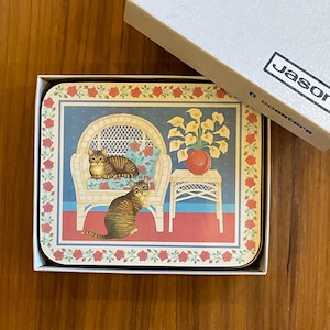 May include: A rectangular Jason coaster set in a white box featuring an illustration of two brown tabby cats, one sitting on a white wicker chair and one on the floor, next to a side table with yellow calla lilies and a red vase, bordered by red roses.