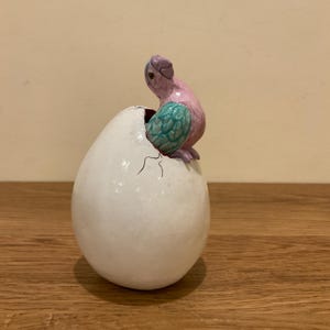 Puede incluir: Figura de cerámica de un pájaro saliendo de un huevo blanco. El pájaro es rosa con un ala turquesa y una cabeza morada. El huevo tiene una superficie agrietada.