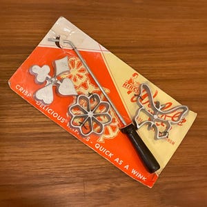 May include: Vintage waffle iron set with three decorative aluminum molds: a flower, a butterfly, and a heart-shaped design. The set includes a long handle with a black grip and is packaged on a red and yellow card.