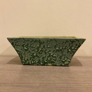 May include: A green ceramic planter with a mottled, speckled finish. The planter has a rectangular shape with slightly flared sides.
