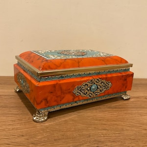 May include: An orange jewelry box with a marble design, gold trim, and turquoise accents. The box has ornate metalwork and rests on four silver-colored feet. The lid features a teal and gold design.