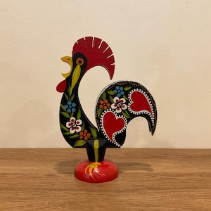 May include: A hand-painted wooden rooster figurine. The rooster is black with red, yellow, and white accents. The body is decorated with floral patterns and heart shapes. The figurine stands on a red base.