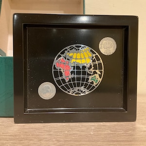 May include: A black wooden box with a glass inset featuring a silver globe with red, yellow, and green continents. The globe is surrounded by three silver coins.