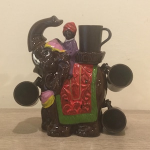 May include: A brown ceramic elephant figurine with a colorful saddle and a small figure of a person riding on top. The elephant has four black cups attached to its sides.