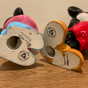 Op de afbeelding: Twee keramische Mickey Mouse-figuurtjes. E&eacute;n draagt een rood shirt en de andere een blauw shirt met een roze vest. Beide figuurtjes hebben zwarte schoenen en zwarte oren. De onderkant van elk figuurtje heeft een wit label met de tekst "Walt Disney Productions Japan" en een logo.