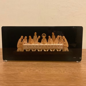 May include: A rectangular, black decorative box featuring a carved relief of the Last Supper. The figures are depicted in a light beige color, with Jesus in the center. The box is set on a wooden surface.