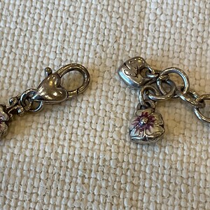 Peut inclure: Un bracelet en m&eacute;tal argent&eacute; dot&eacute; d'un fermoir mousqueton en forme de c&oelig;ur et d'une petite breloque en forme de fleur aux p&eacute;tales blancs avec un centre violet. La cha&icirc;ne est compos&eacute;e de maillons circulaires et inclut une breloque en c&oelig;ur.
