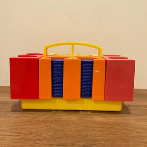 May include: A rectangular plastic game storage case with a yellow base and handle, featuring red and orange vertical dividers. Two central compartments are filled with stacks of small, round, dark blue game tokens or chips.