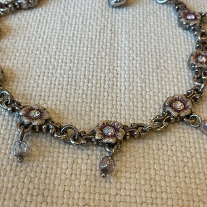 Peut inclure: Un bracelet &agrave; breloques en m&eacute;tal argent&eacute; compos&eacute; de maillons en forme de fleurs avec des d&eacute;tails en &eacute;mail violet et des cristaux transparents au centre. De petites perles rondes violettes translucides pendent des maillons floraux.
