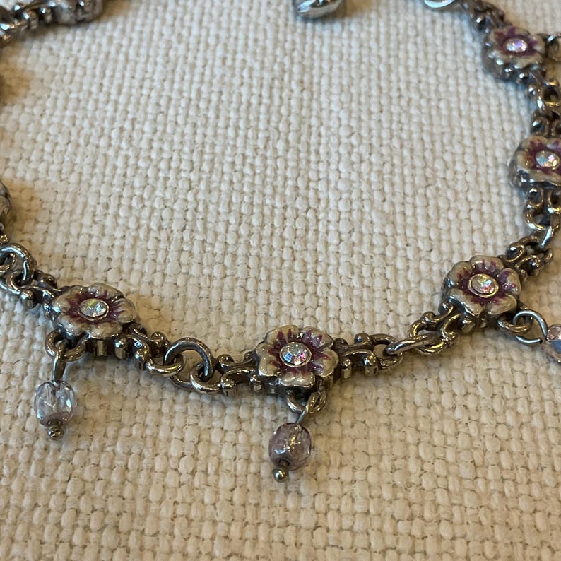 Peut inclure: Un bracelet &agrave; breloques en m&eacute;tal argent&eacute; compos&eacute; de maillons en forme de fleurs avec des d&eacute;tails en &eacute;mail violet et des cristaux transparents au centre. De petites perles rondes violettes translucides pendent des maillons floraux.