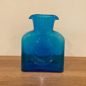 May include: A vibrant, turquoise-blue glass vase with a square base and a flared neck. The vase has a rounded body and a smooth, glossy finish. The vase is empty and sits on a wooden surface.
