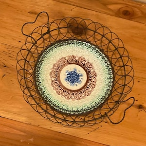 May include: A decorative plate with a floral design in a metal wire basket. The plate features a blue, brown, and green color scheme. The basket has two heart-shaped handles.