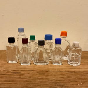 May include: A collection of eight small, clear glass bottles of varying shapes and sizes, each topped with a different colored plastic cap including black, light blue, maroon, green, dark blue, orange, and white, arranged in a group.
