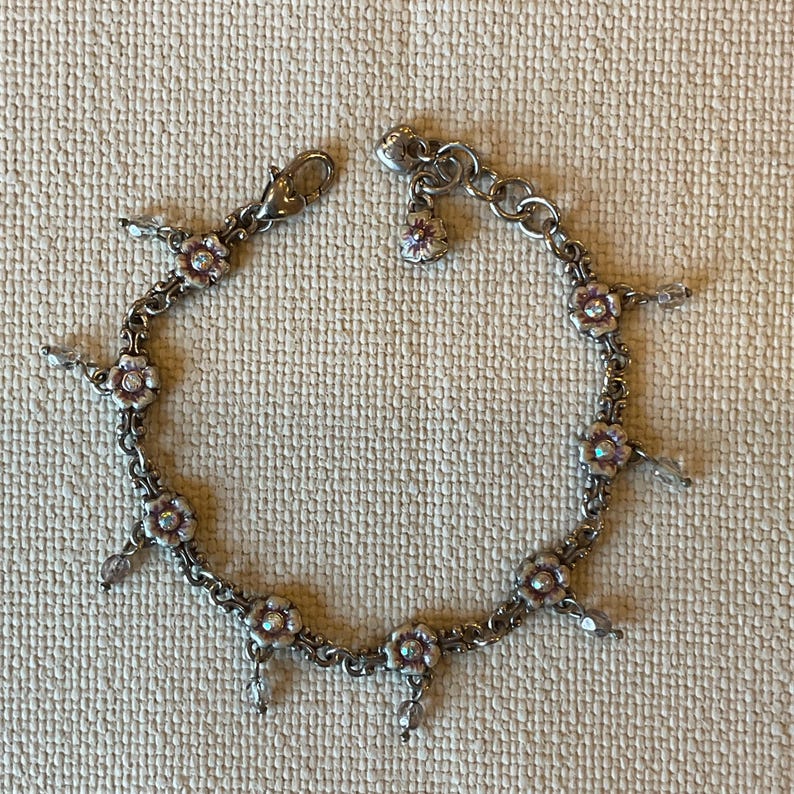 Peut inclure: Un bracelet &agrave; breloques argent&eacute; compos&eacute; d'une s&eacute;rie de petites fleurs &agrave; cinq p&eacute;tales avec des centres iris&eacute;s. Le bracelet comprend un fermoir mousqueton, une petite breloque en forme de c&oelig;ur et des perles de cristal transparentes.