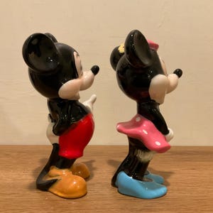 Op de afbeelding: Een paar keramische beeldjes van Mickey Mouse en Minnie Mouse. Mickey draagt een rode broek en gele schoenen. Minnie draagt een roze gebloemde jurk en blauwe schoenen.