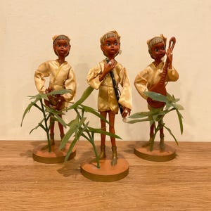 May include: Three vintage dolls with dark skin tones, wearing gold-colored outfits and headbands. Each doll is posed with a different accessory, including a musical instrument and a purse. The dolls are mounted on round, tan bases with green plant accents.