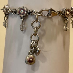 Peut inclure: Un bracelet &agrave; breloques argent&eacute; orn&eacute; de motifs floraux r&eacute;p&eacute;titifs avec des centres iris&eacute;s. Le bracelet comprend des perles transparentes pendantes et un groupe de breloques central compos&eacute; d'une petite fleur en m&eacute;tal et d'un c&oelig;ur sur une cha&icirc;ne.
