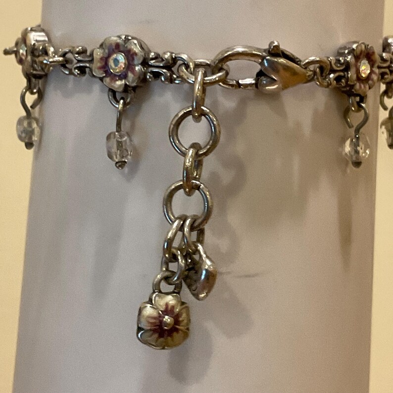 Peut inclure: Un bracelet &agrave; breloques argent&eacute; orn&eacute; de motifs floraux r&eacute;p&eacute;titifs avec des centres iris&eacute;s. Le bracelet comprend des perles transparentes pendantes et un groupe de breloques central compos&eacute; d'une petite fleur en m&eacute;tal et d'un c&oelig;ur sur une cha&icirc;ne.