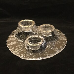 May include: A clear glass candle holder with three candle cups. The holder has a textured, frosted surface and a round base.