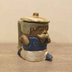May include: A ceramic mug shaped like a man wearing a blue shirt, white pants, and a hat. The mug has a lid and a handle.