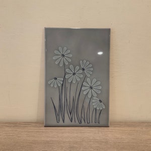 May include: A gray ceramic tile with a design of white flowers and black stems on a gray background.
