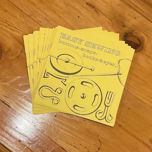 May include: A stack of yellow paperbacks with the title "EASY SEWING buttons, snaps, hooks & eyes." The cover features a black and white illustration of buttons, snaps, hooks, and eyes.