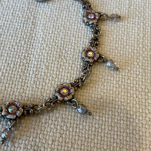 Peut inclure: Un collier &agrave; cha&icirc;ne en m&eacute;tal argent&eacute; compos&eacute; de maillons en forme de fleurs avec des d&eacute;tails en &eacute;mail violet et un strass transparent au centre. De petites perles transparentes &agrave; facettes sont suspendues entre les segments floraux.